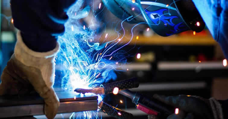 Smoke and sparks are being transmitted during a welding application. A welder in a protective mask is holding the workpiece with one hand and welding with the other.