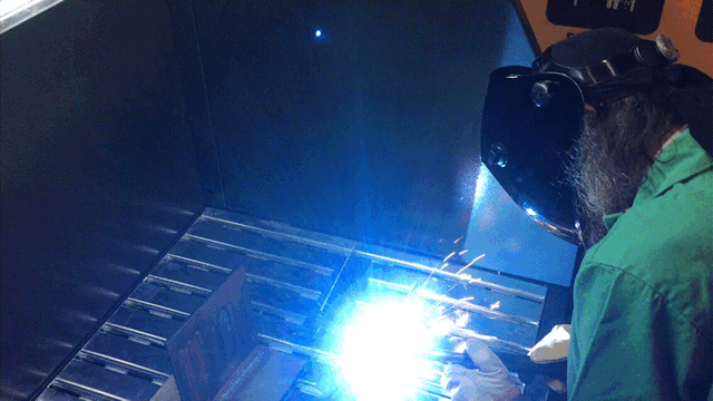 Weld fumes and sparks are being sucked down into a downdraft table. A machine operator in protective wear is performing a welding application on top of the downdraft table.