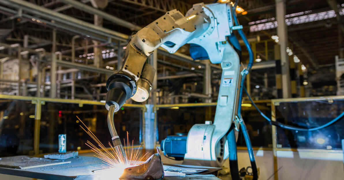 A robotic welding machine in the process of a work piece. Robotic welding sparks being emitted from the workpiece and a manufacturing environment in the near background.